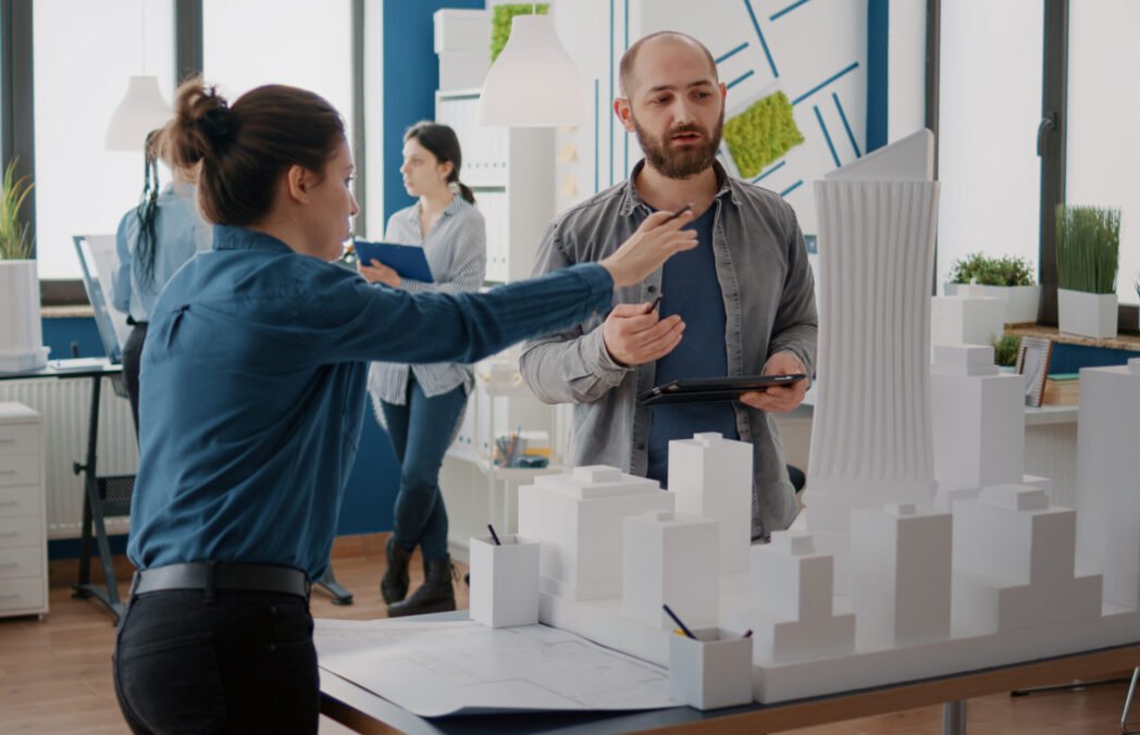 Team of coworkers analyzing maquette and building model to plan construction on tablet. Architectural people working together, designing urban strategy and real estate development.