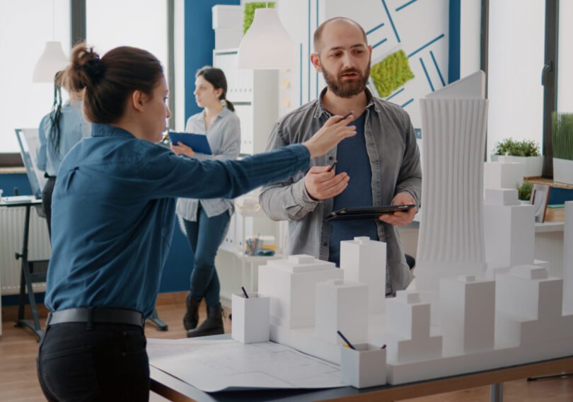 Team of coworkers analyzing maquette and building model to plan construction on tablet. Architectural people working together, designing urban strategy and real estate development.